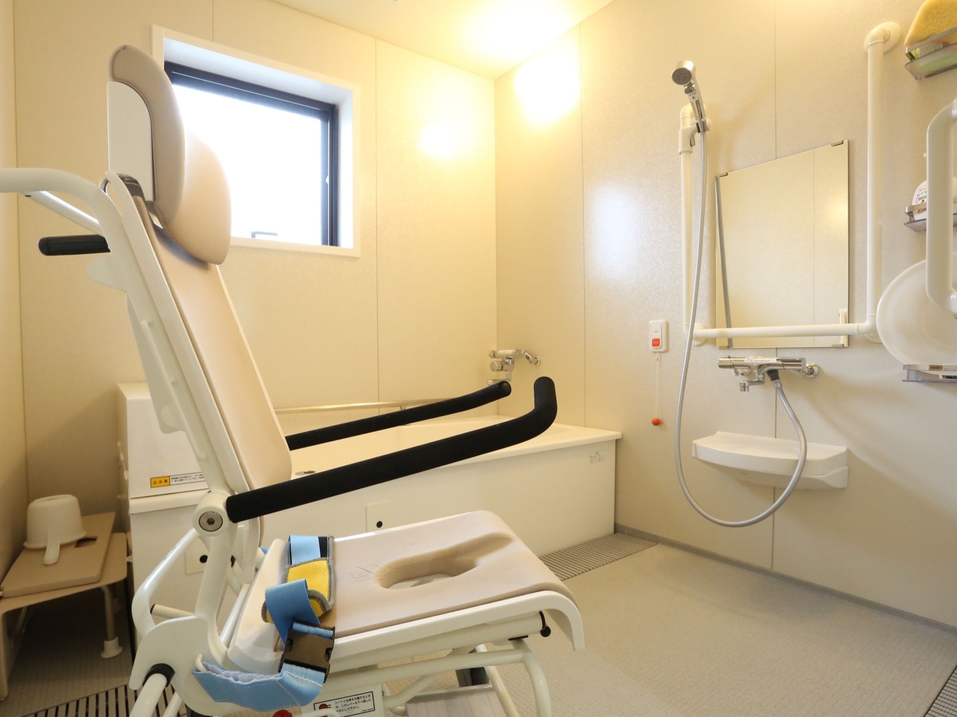 白を基調とした清潔感のある浴室。入浴介助用のシャワーチェアが置かれている。鏡の横に手すりが設置されている。