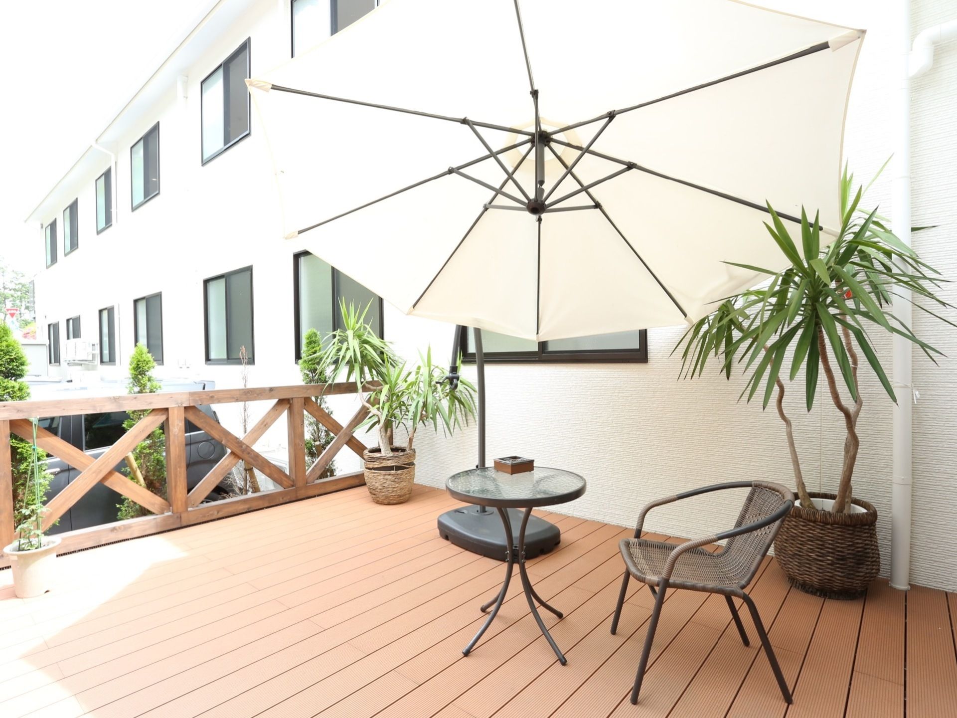 ダークブラウンの木を基調としたベランダに、観葉植物が置かれている。日よけのパラソルと１人用のテーブル、椅子が設置されている。