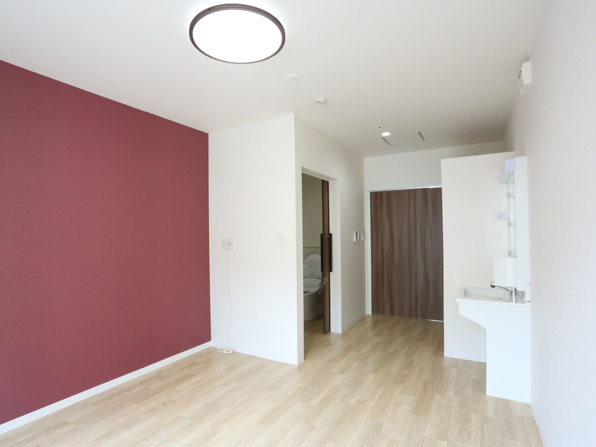 木目調の床にボルドー色の壁紙が映えている。洗面台が置かれており、室内に清潔感のあるトイレが備わっている。