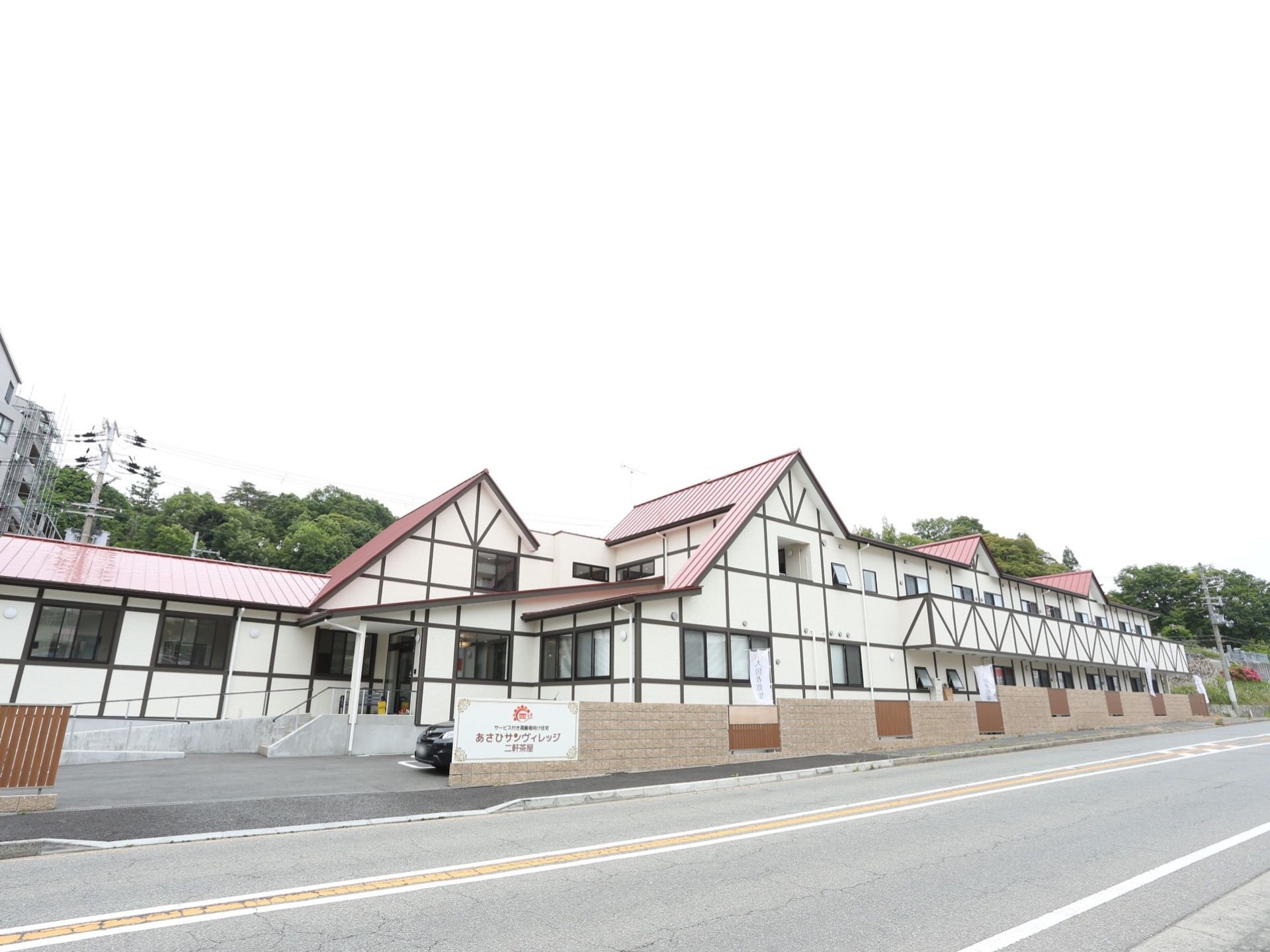 白の外壁に黒のラインが映える二階建ての施設。施設の周りにはピンクベージュの塀が建てられており、塀に施設名の看板が設置されている。