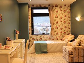 花柄のクロスが印象的な明るい部屋。寝具付きのベッドがあり、一人用の椅子やデスクセットも置かれている。