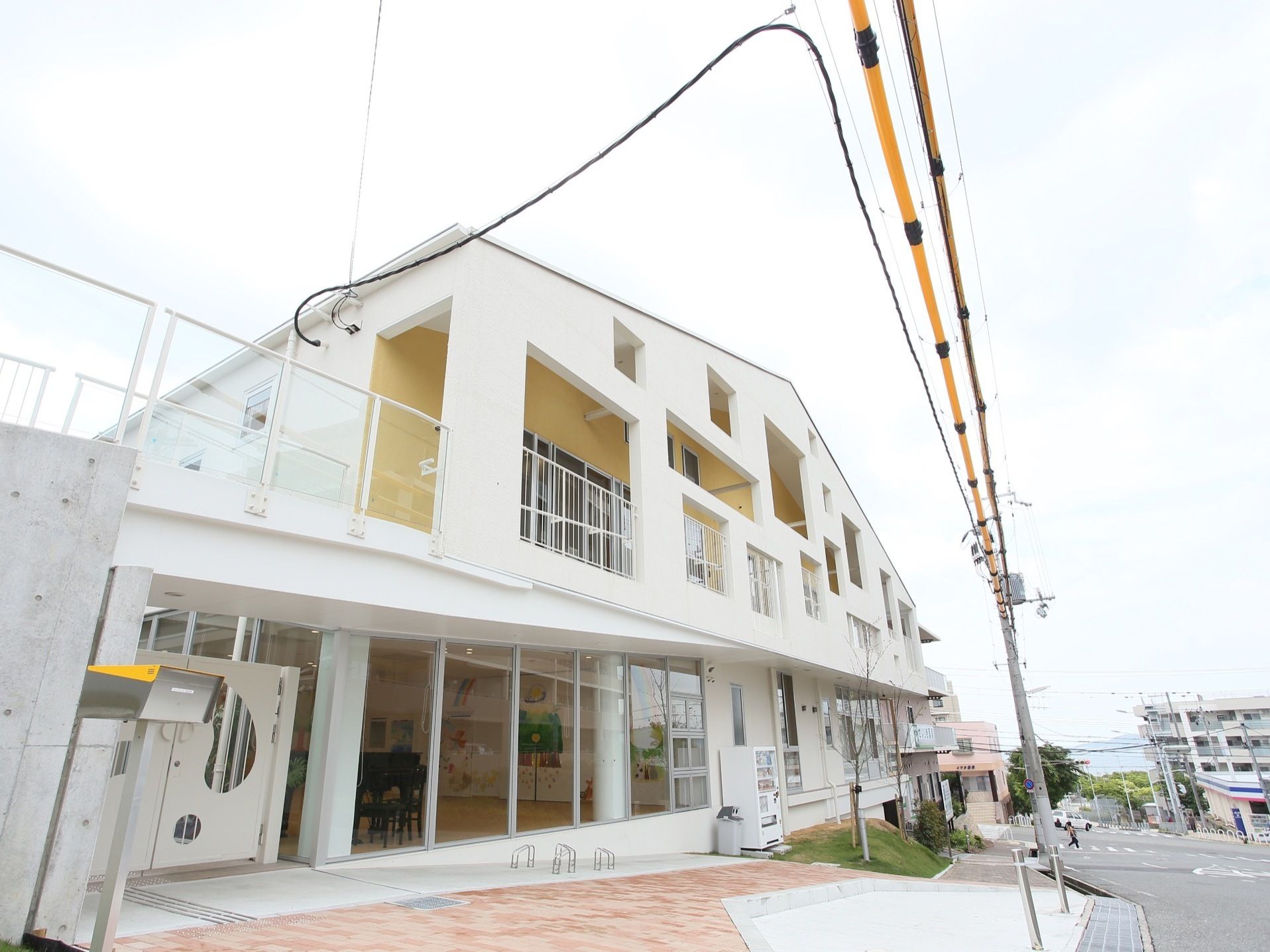 3階建ての白い建物がある。入口までは緩やかな坂になっていて、建物の壁沿いに自動販売機が設置されている。