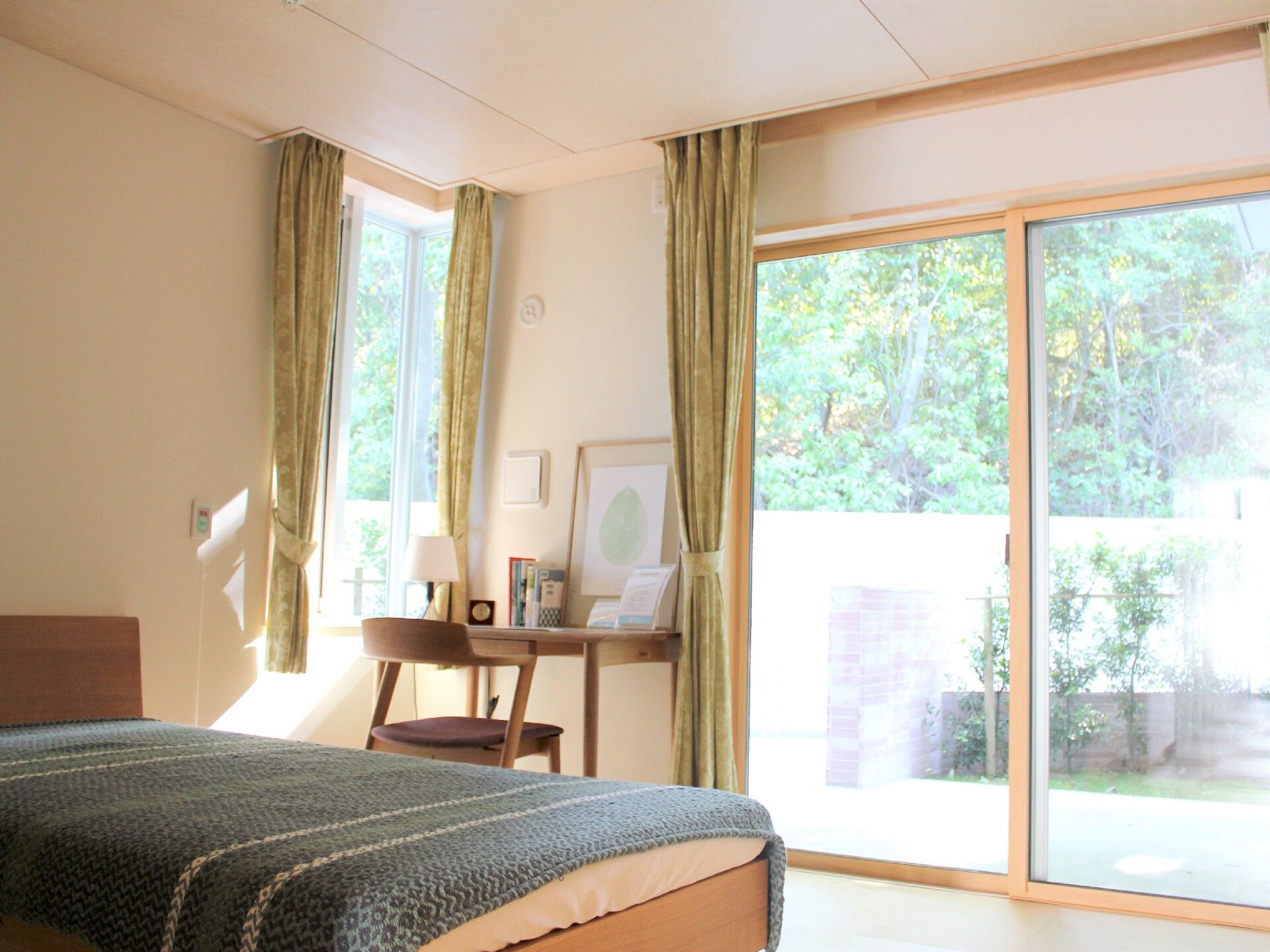 周辺の緑を見ることができる大きな窓が付いたフローリングの部屋には緑のカバーがつけられた寝具付きのベッドがあり、デスクセットにはランプも置かれている。