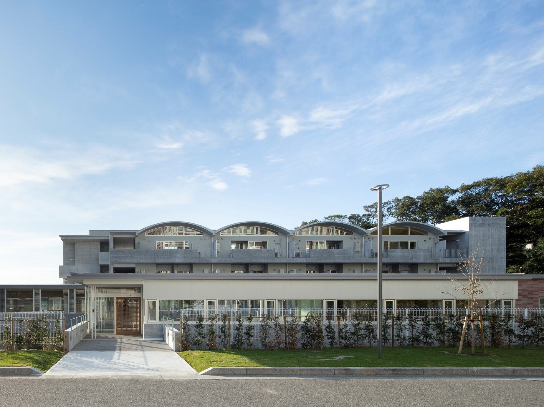 半円状の屋根が連なっている白い建物の周りには緑が多く植えられ、芝生スペースや玄関に続く幅の広い白いアプローチもある。