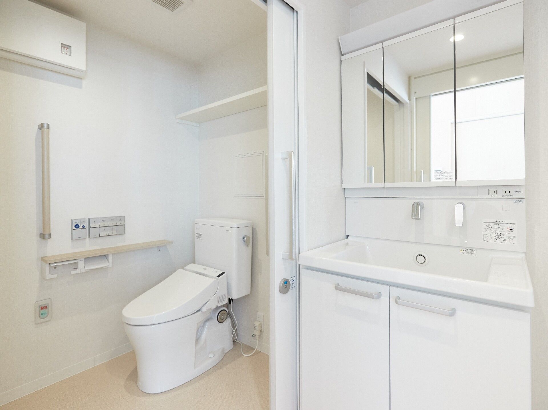 左側には手すりや呼び出しボタンが付いたトイレがあり、便座の上には棚がある。右側には収納や鏡が付いた洗面所がある。