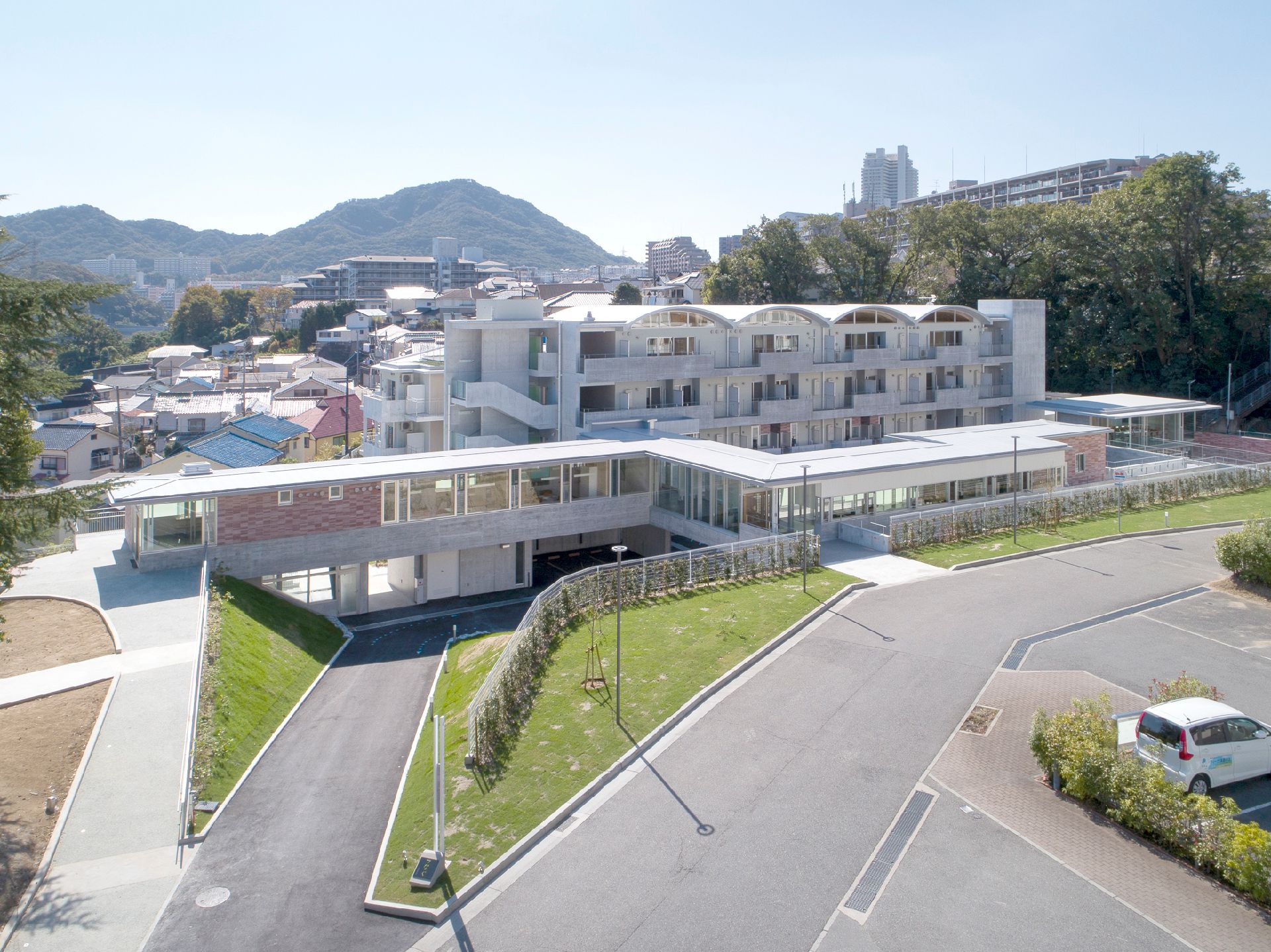 緑に囲まれた場所にあり、中央に白い建物が建っている。手前には駐車場があり、奥には山々が連なっているのが見える。