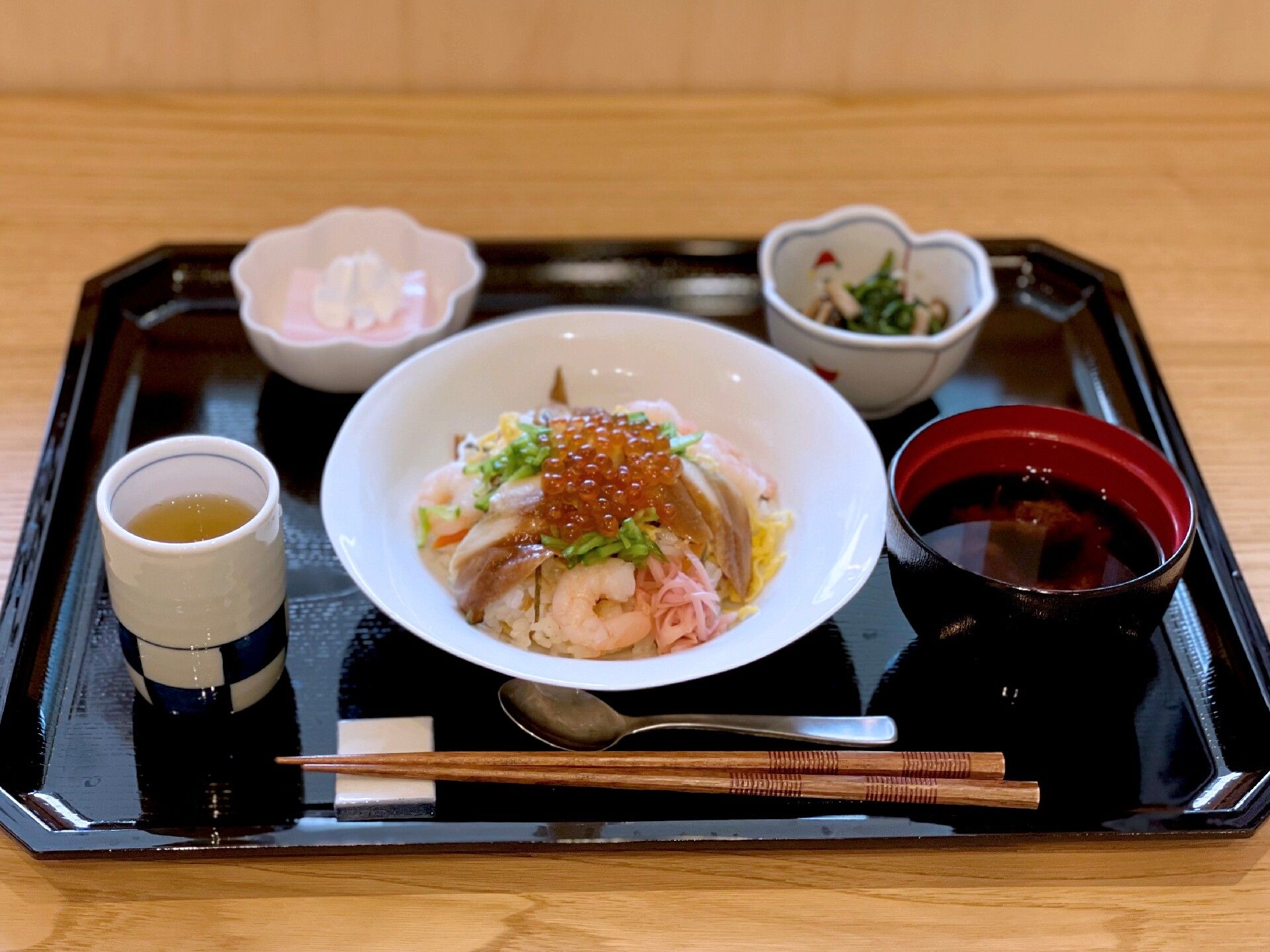 イクラやエビ、錦糸卵などが乗った丼が中央に置かれ、お茶や汁物、子×などが並んだ黒いトレーには箸も添えられている。