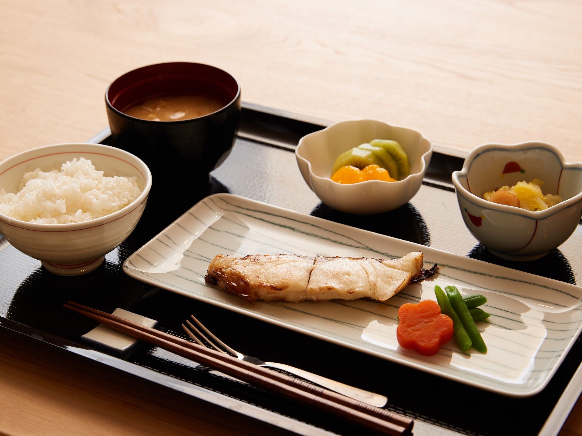 長細い冬季の皿には焼き鮭と人参、いんげんが盛られ、白いご飯にみそ汁、野菜の小鉢などが黒いトレーに並べられている。