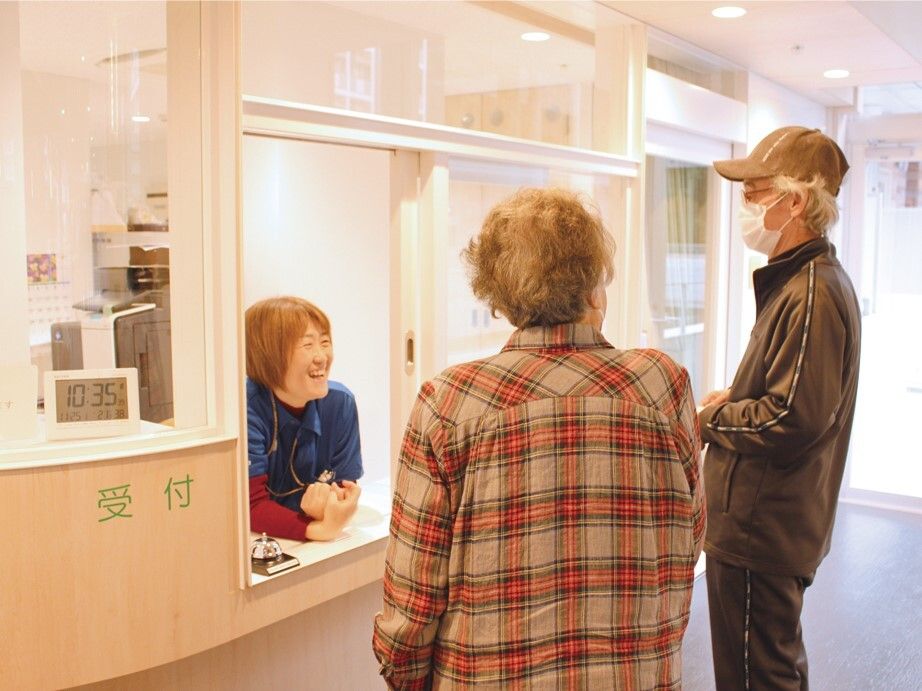 明るい笑顔で対応している女性スタッフが受付にいて、その前にはマスクを着け帽子をかぶった高齢男性と高齢女性がいる。