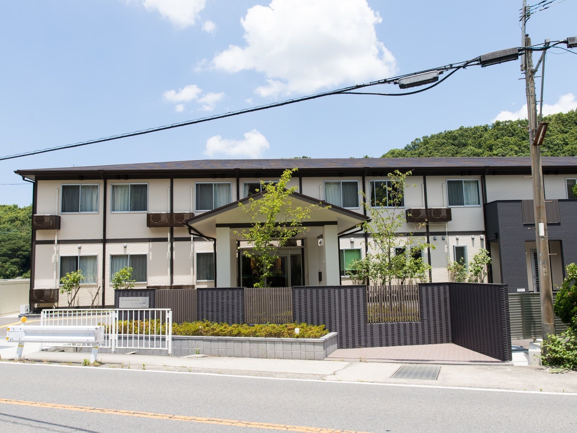 緑に囲まれた環境に立地する施設である。目の前は車道が走っている。電柱が立ってある。緑地も完備されている。