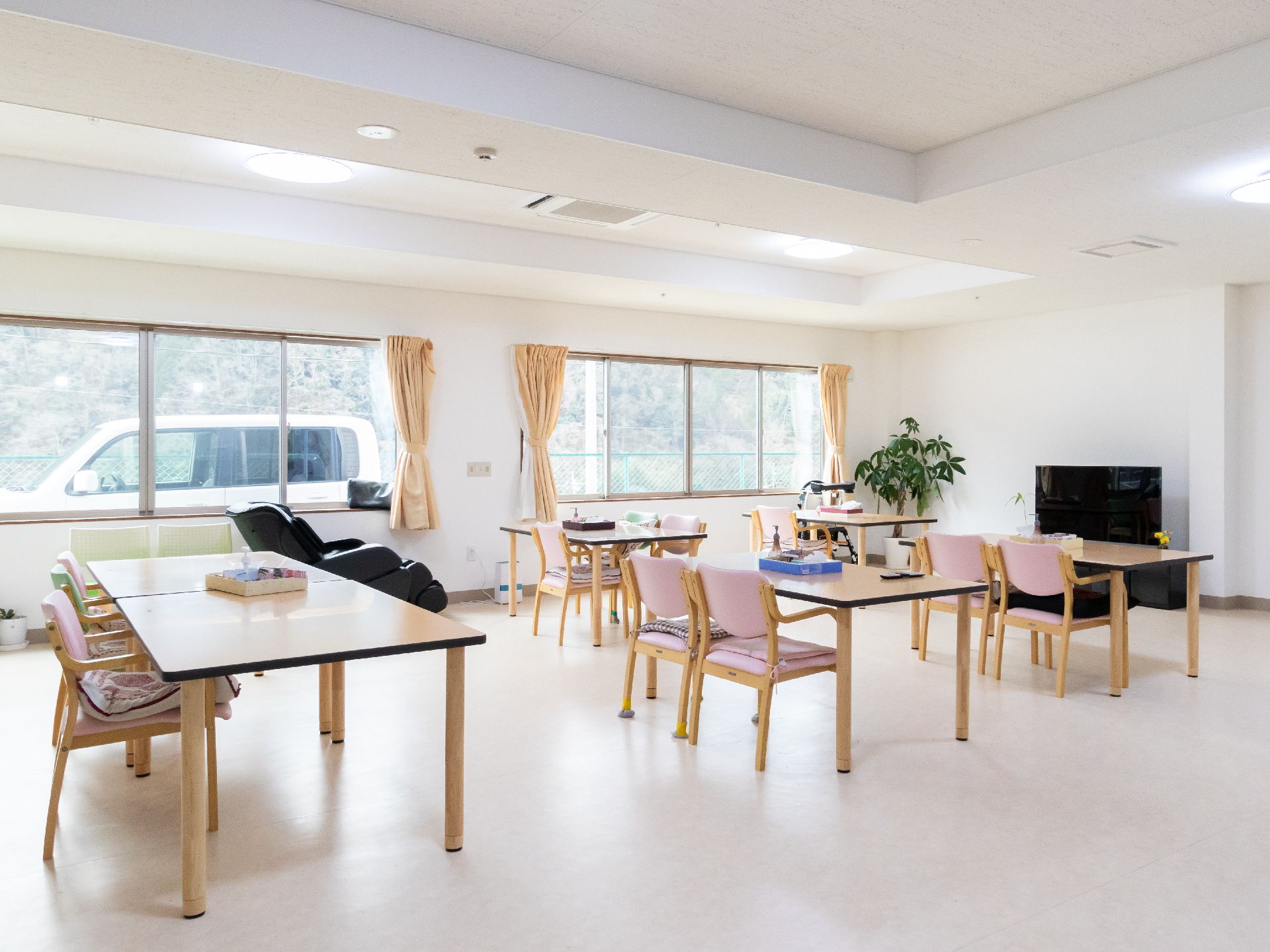 複数のテーブルとピンク色の介護用椅子が並べられた液晶テレビがある広間