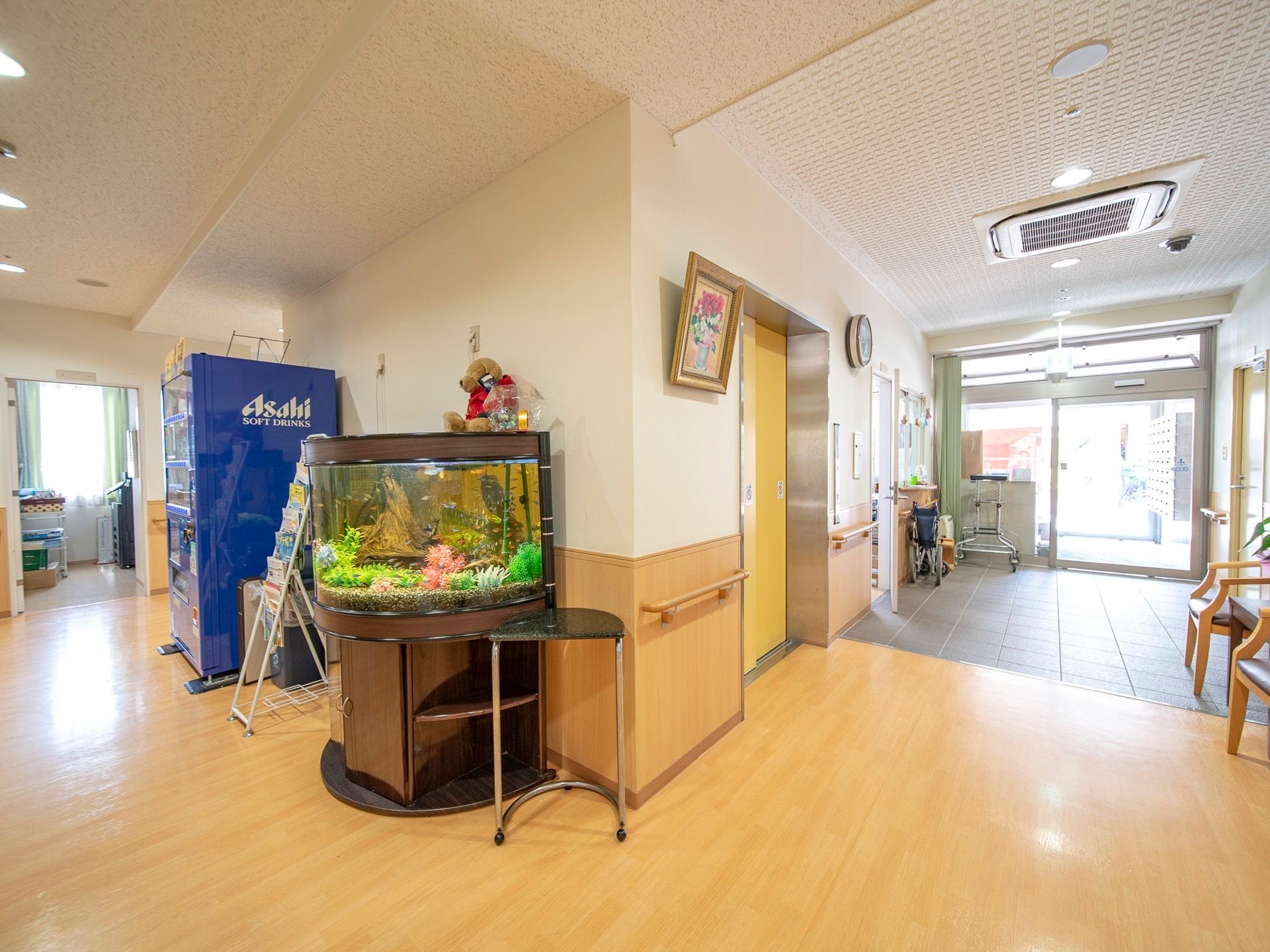 透明なガラス戸の玄関。通路を曲がったところに、半円柱型の水槽とぬいぐるみが飾られており、その奧に自動販売機が置かれている。