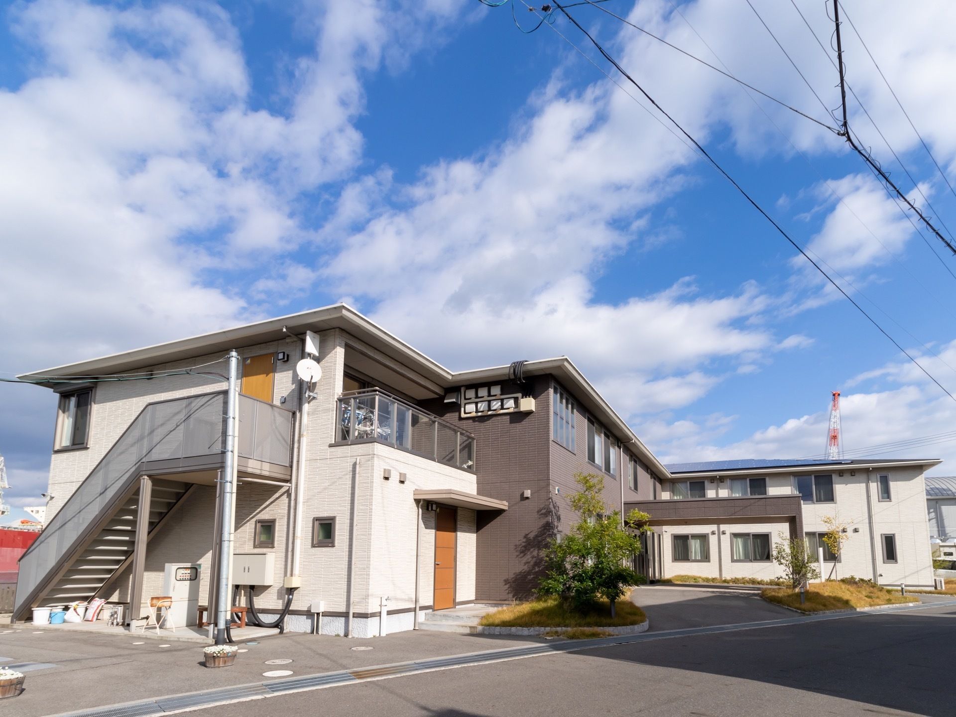白とグレーの外壁の２階建ての施設である。建物前に芝生スペースがあり、そこに高さのある樹木が植えられている。