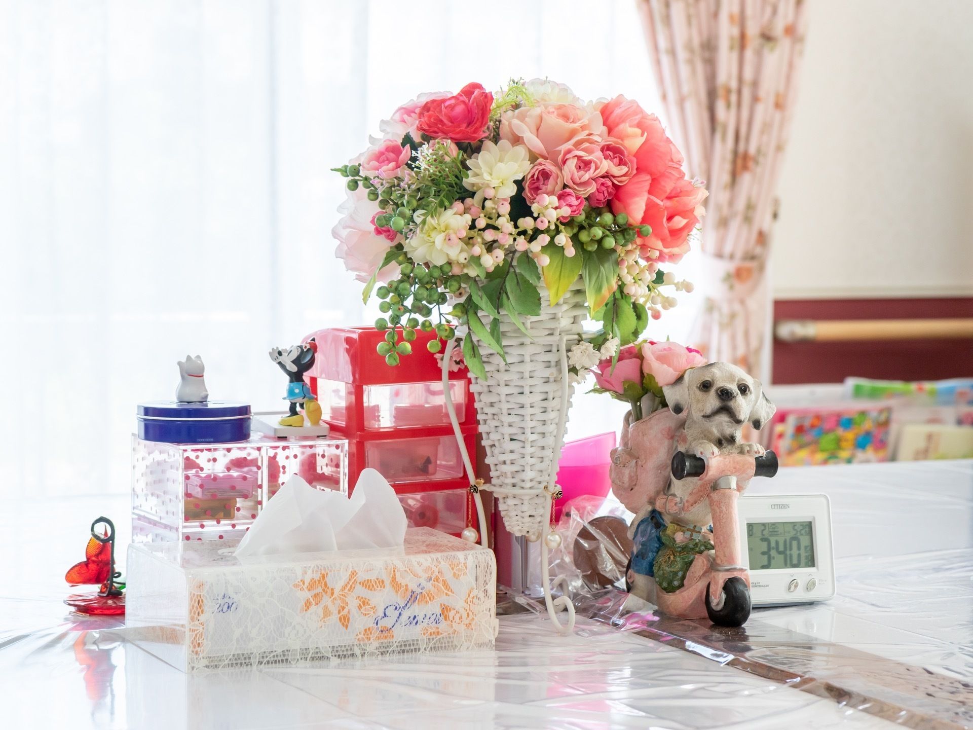 細かい模様のピンク色のカーテンが掛けられた部屋である。テーブルに白いかごの花のアレンジメントが飾られ、時計が置いてある。
