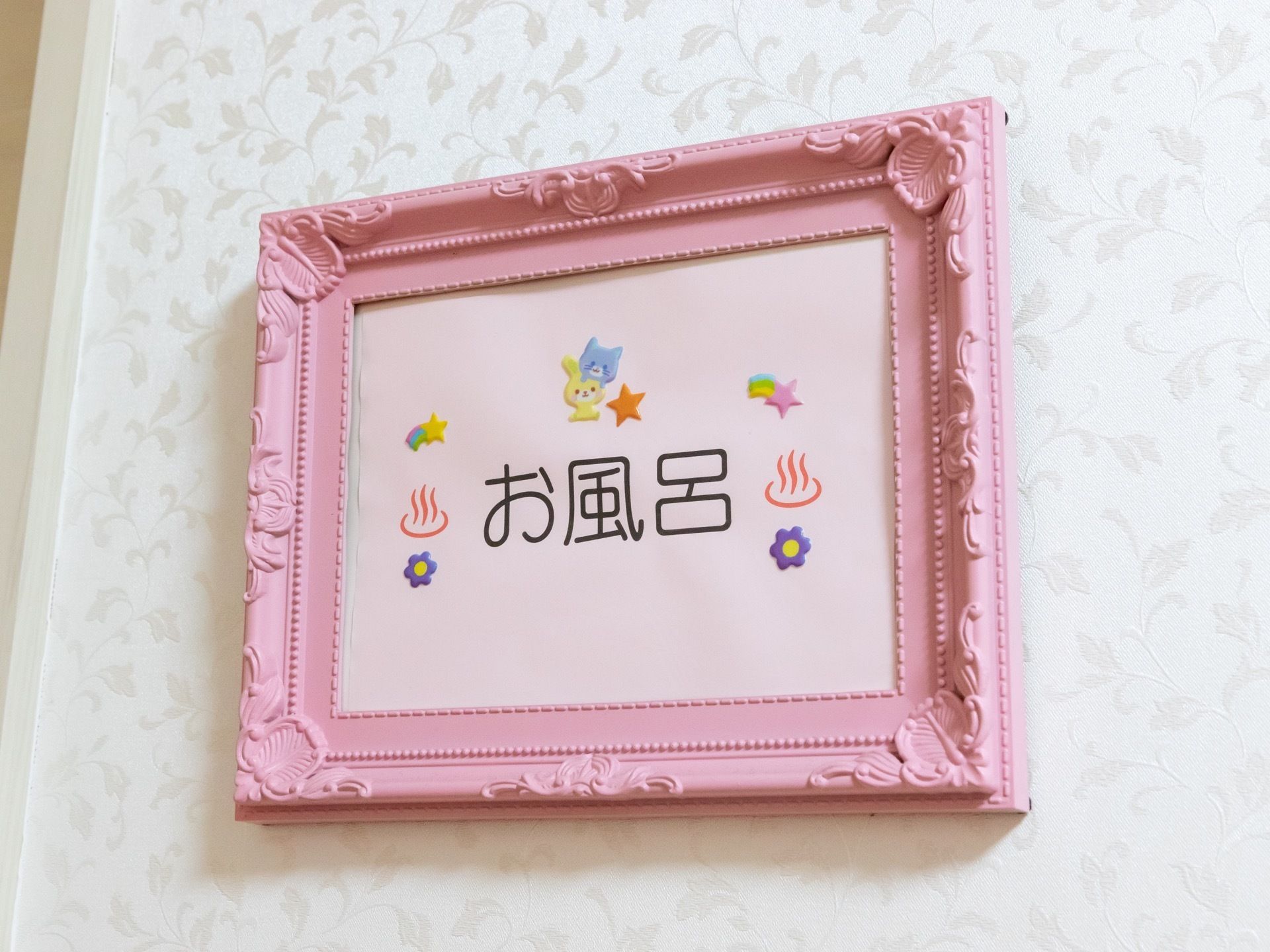 浴室を示す案内坂。お風呂の文字と猫や星のイラストの入った紙が、ピンクの額縁に入れられ、白い壁に掛けられている。