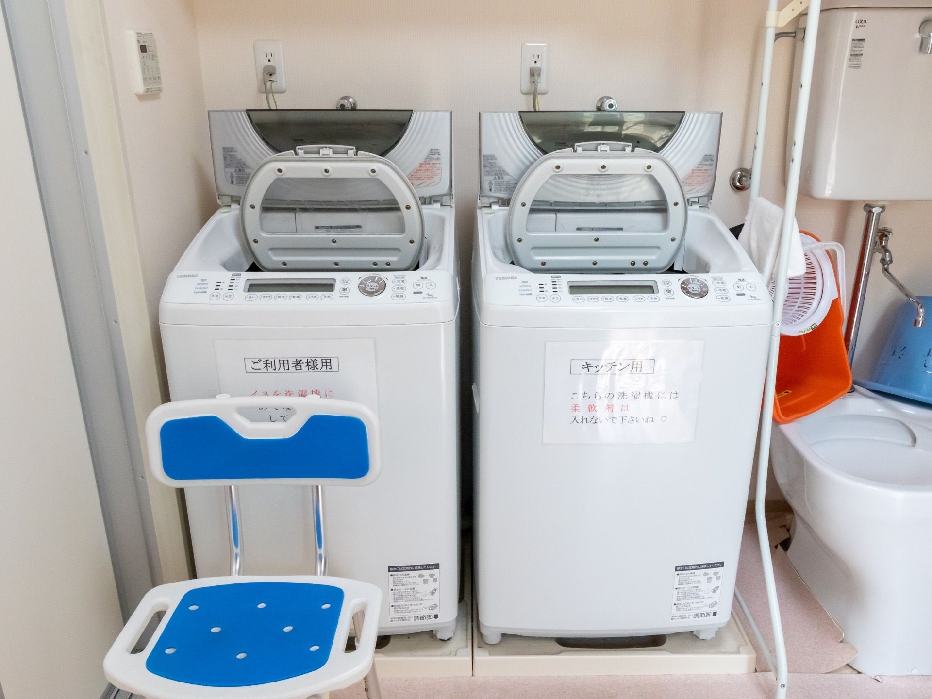 ２台の縦型の洗濯機が並べられ、蓋が開けられている。高い位置にタンクが設置された汚物流しと青い椅子が置いてある。