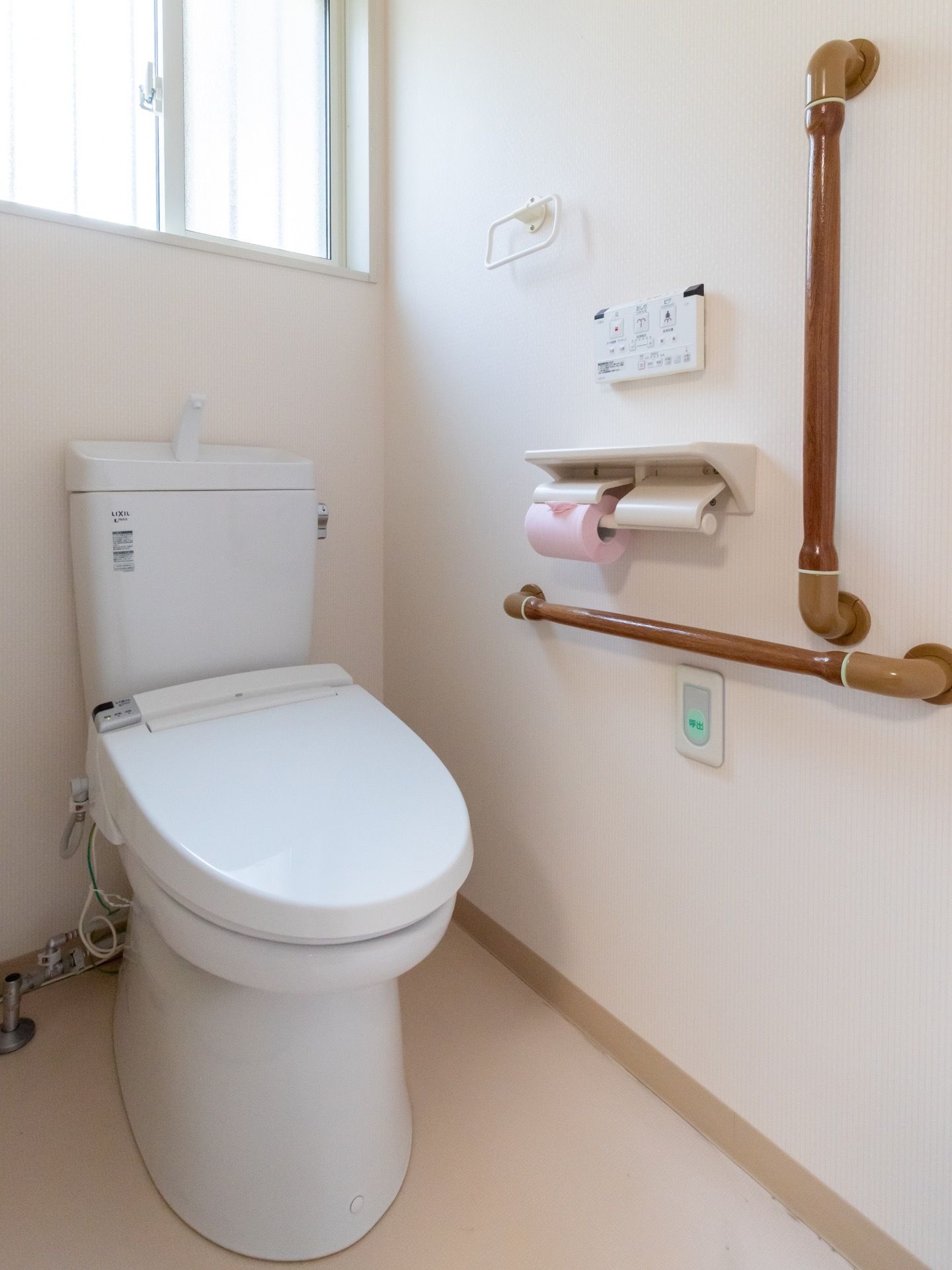 手洗いタンク付きの白い洋式トイレがある。壁に手すりとリモコンとペーパーホルダーが設置され、ピンク色のトイレットペーパーが取り付けられている。