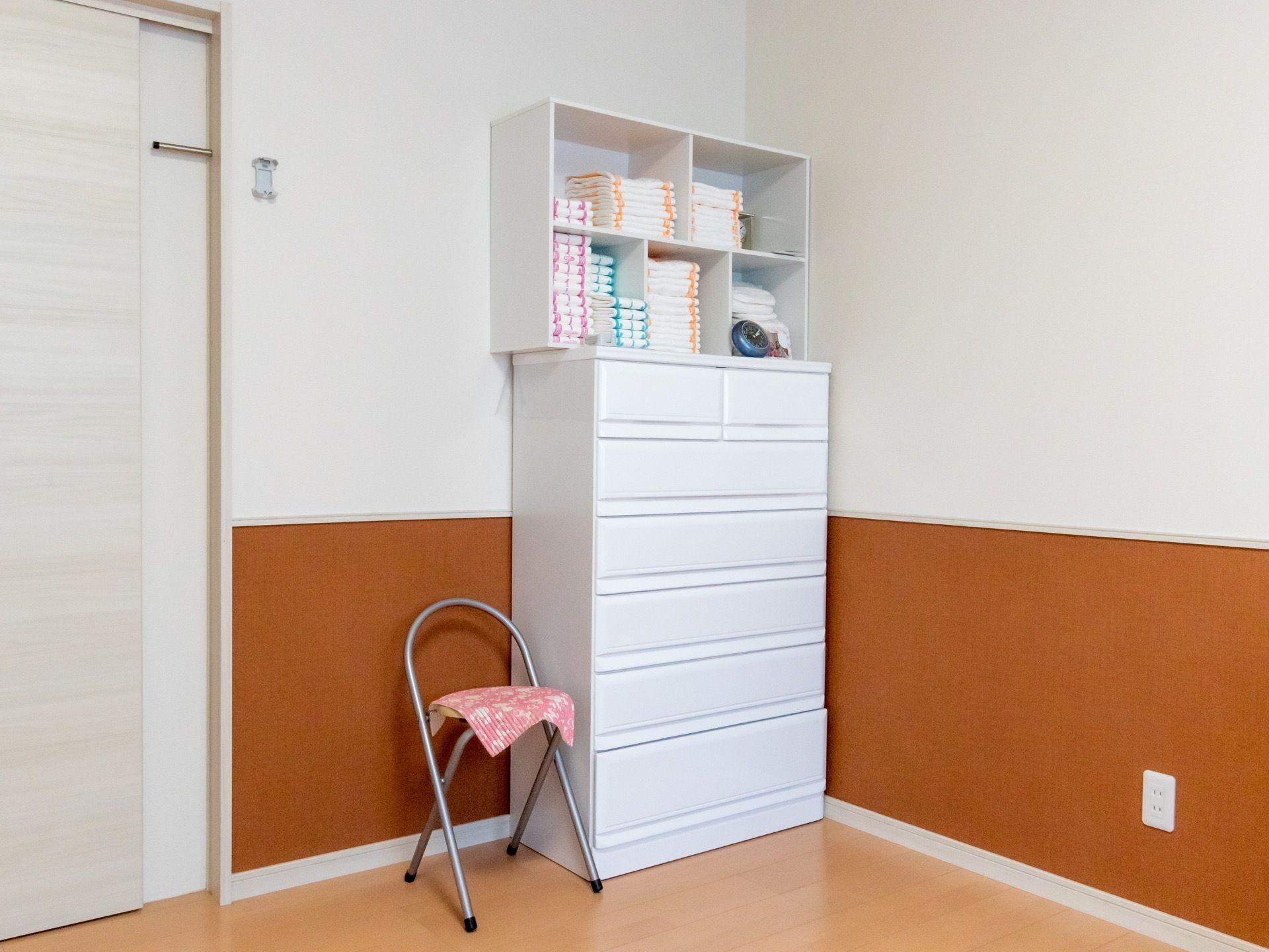 白を基調とした明るい居室である。壁の色にマッチした洋服タンスや椅子が置かれている。棚にはタオルがたくさん収納されている。