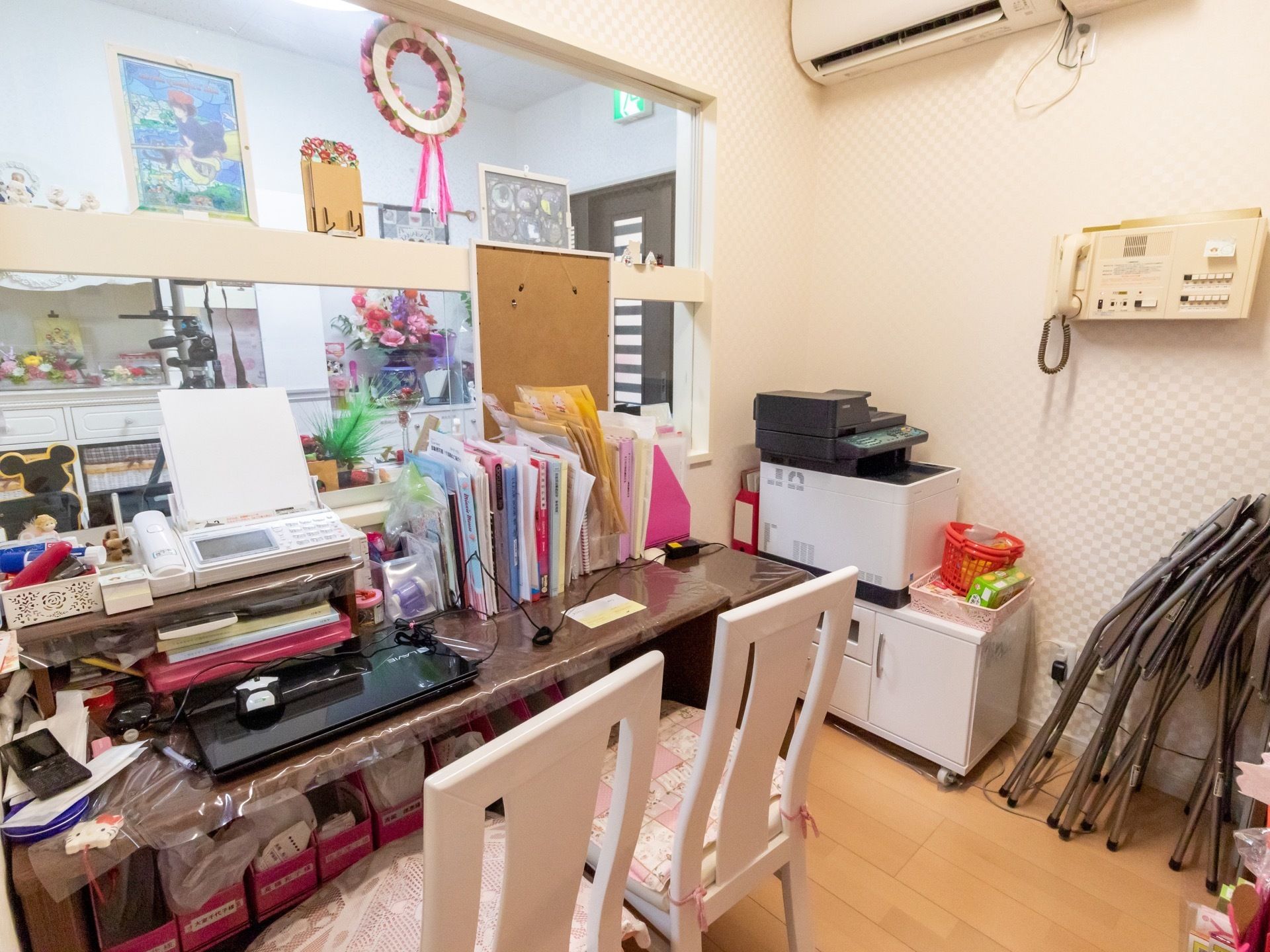 テーブルの上にたくさんの書類が立てかけられ、椅子が２脚並べて置いてある。壁に電話機やエアコンが設置されている。