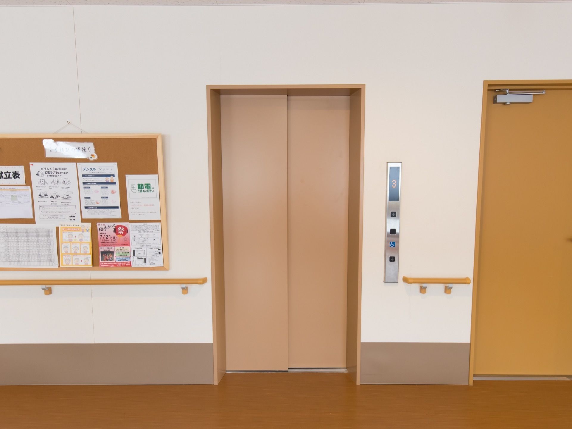 通路の一角にエレベーターが設置されている。扉の左側の壁面には、掲示物が取り付けられたコルクボードが展示してある。
