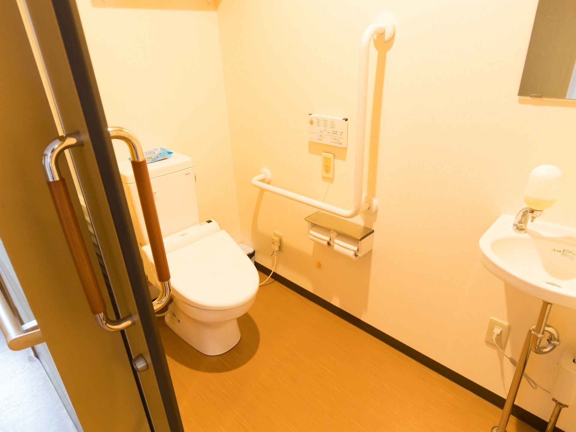 引き戸の奥に蓋の付いた洋式便器が設置されている。手すりのそばに、トイレ操作や非常時にスタッフを呼び出すためのボタンが取り付けてある。