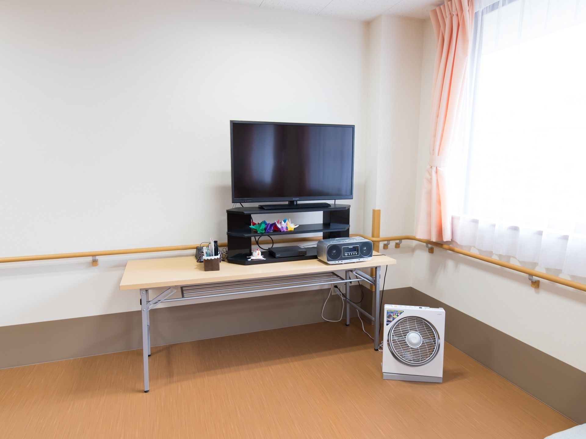 壁面に手すりが取り付けられている部屋の隅にテーブルが置かれている。その上にテレビ台と液晶テレビが載せてある。