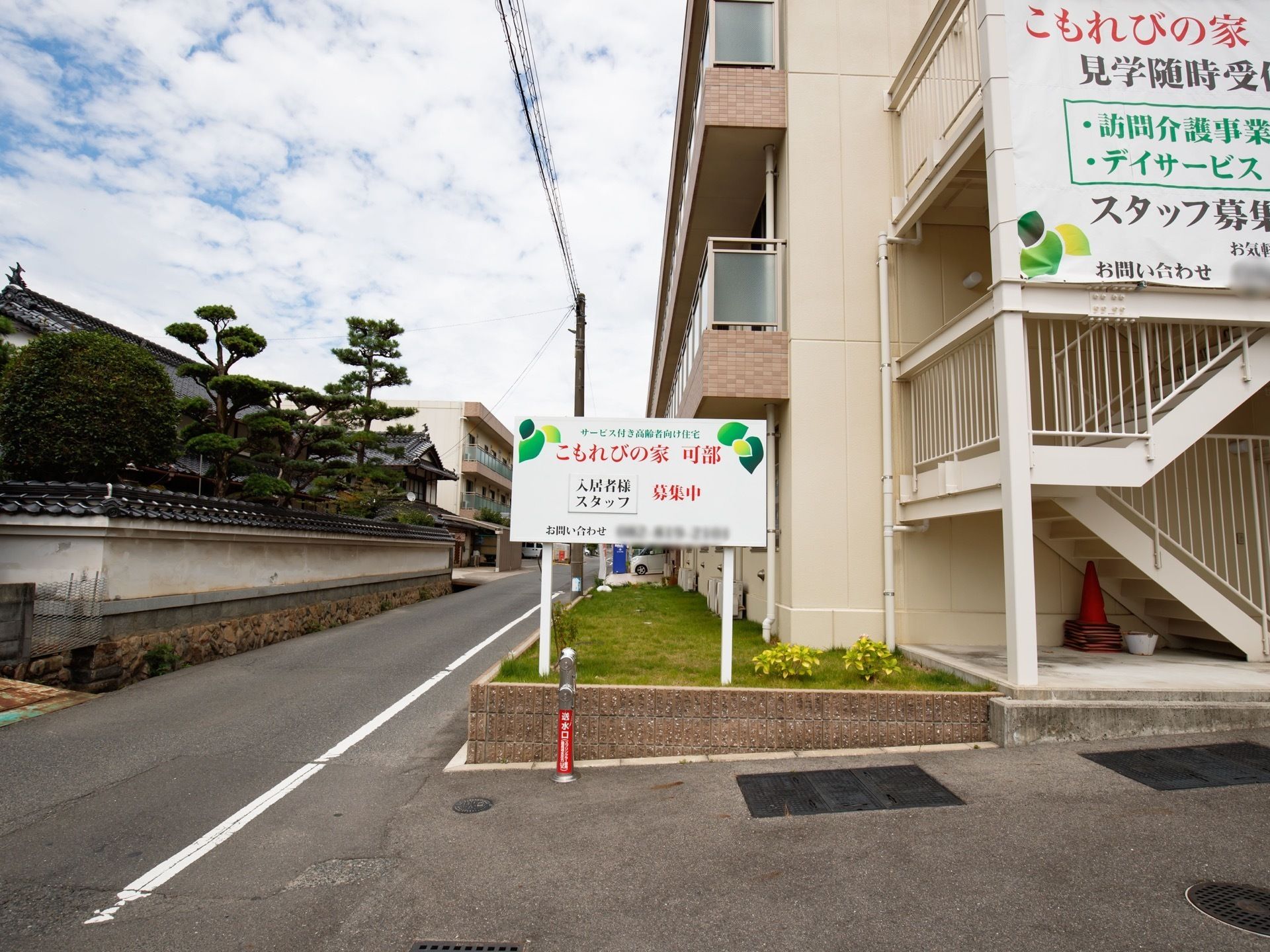 建物の外階段に横断幕が設置されており、施設の説明が書かれている。道路の脇にある芝生スペースに、施設名称が書かれた看板がある。