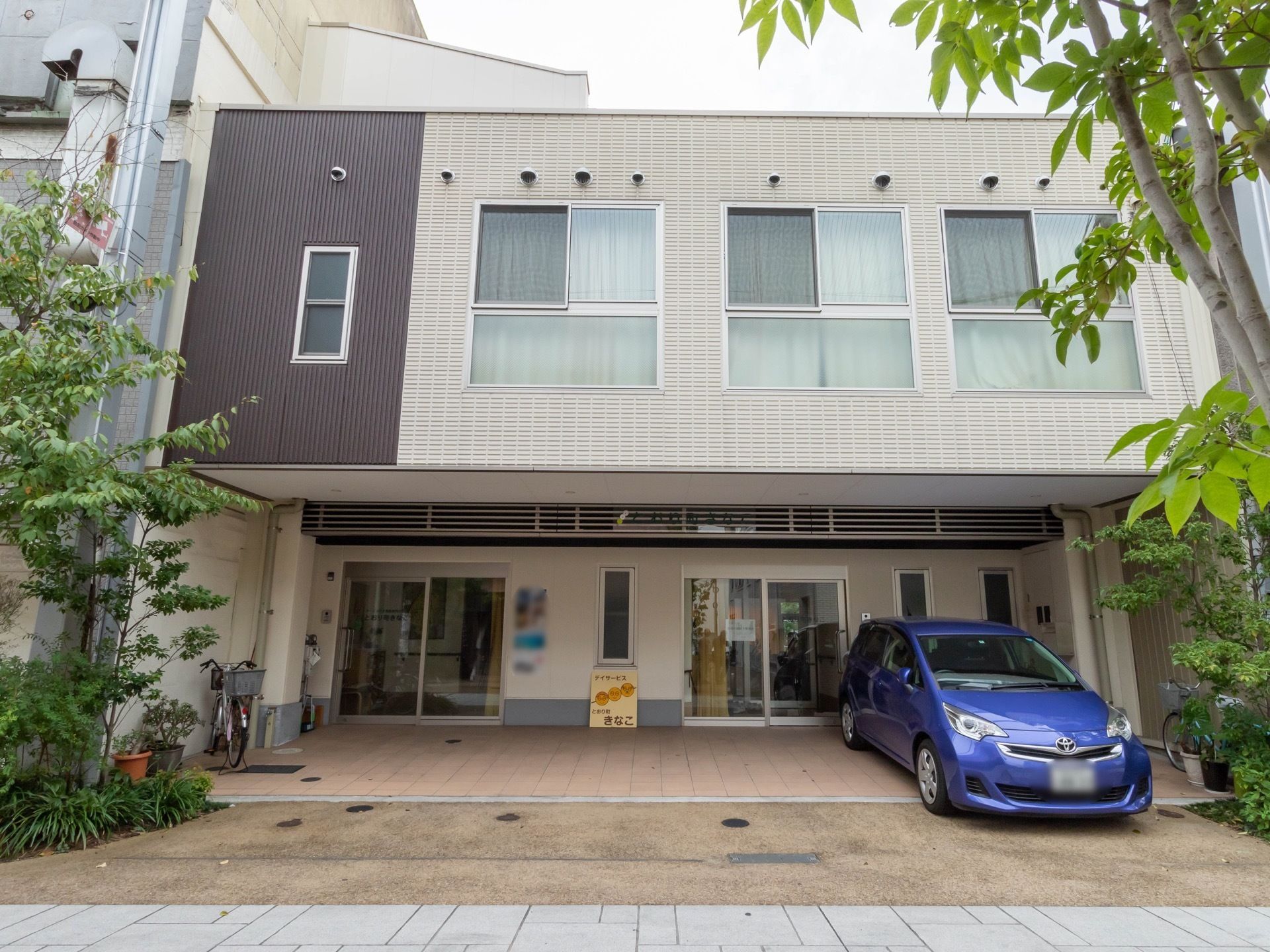 玄関部分が屋根付きのスペースになった２階建ての住宅がある。道路から玄関まで段差のない造りで、青い車が一台停められている。