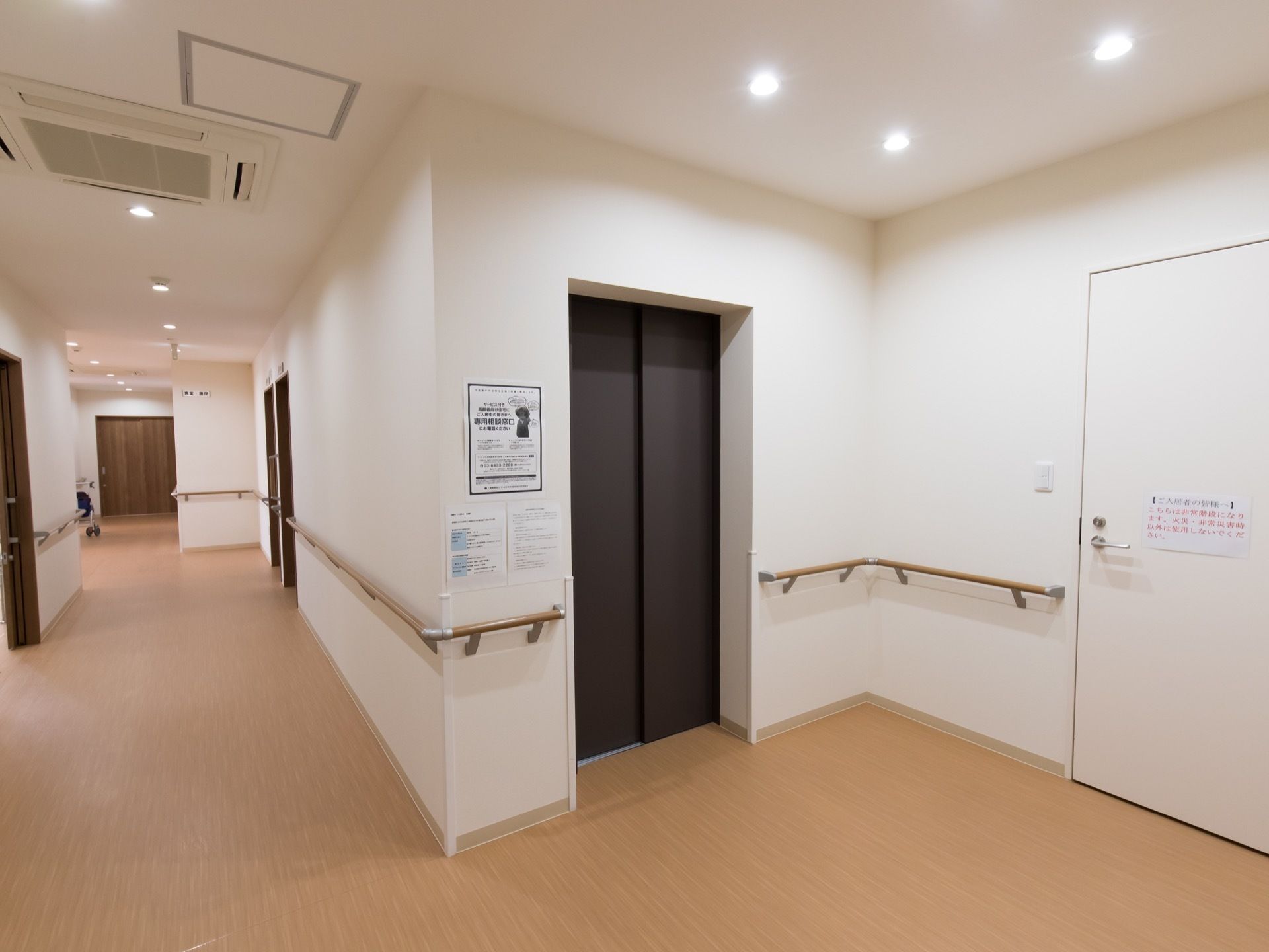 清掃の行き届いた明るい通路。壁面には手すりが取り付けられており、黒い扉のエレベーターが設置されている。