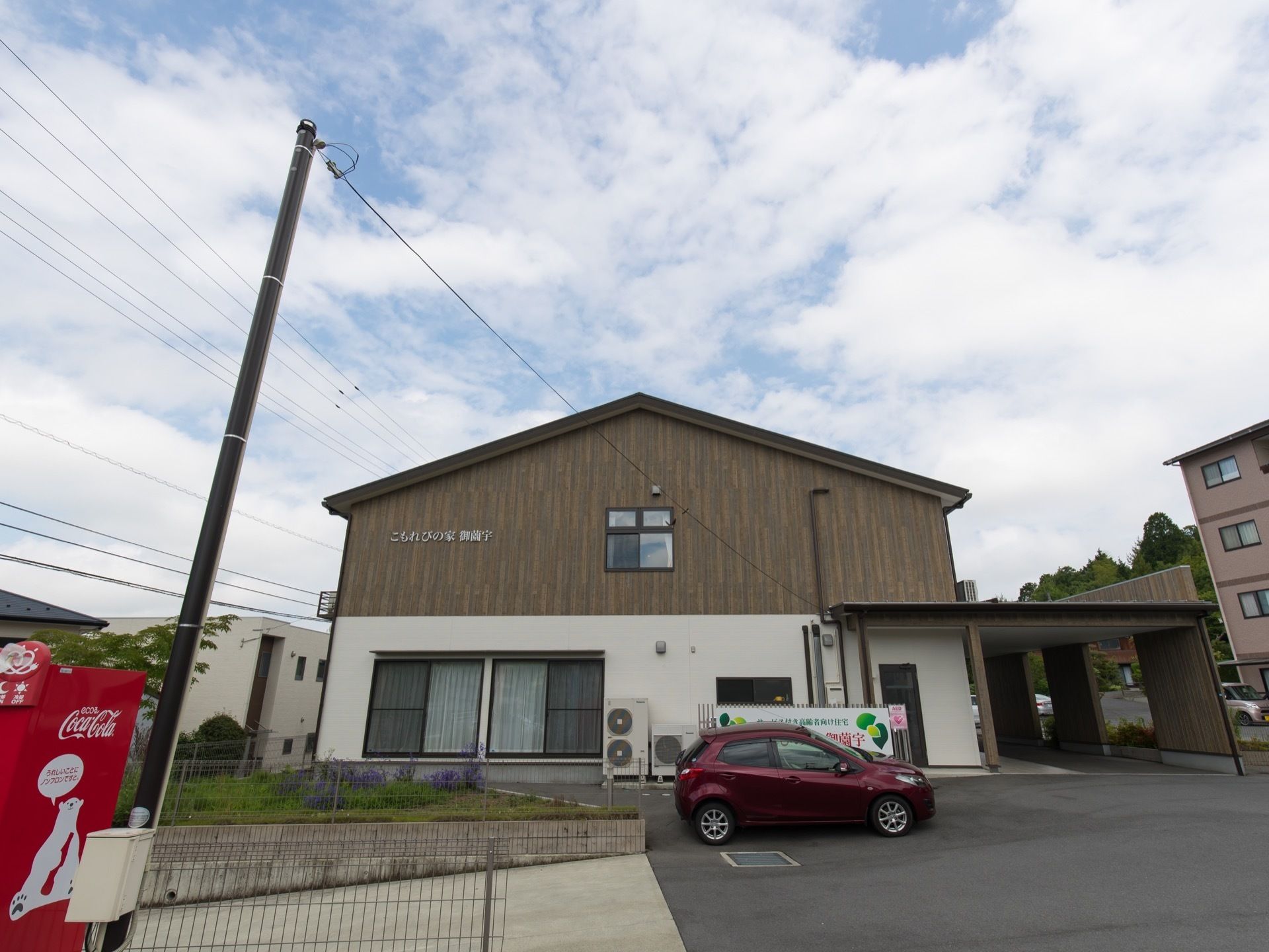 舗装され植栽が施された敷地に、木目調の外壁が美しい２階建ての施設が建っている。手前に飲料の自動販売機が見える。