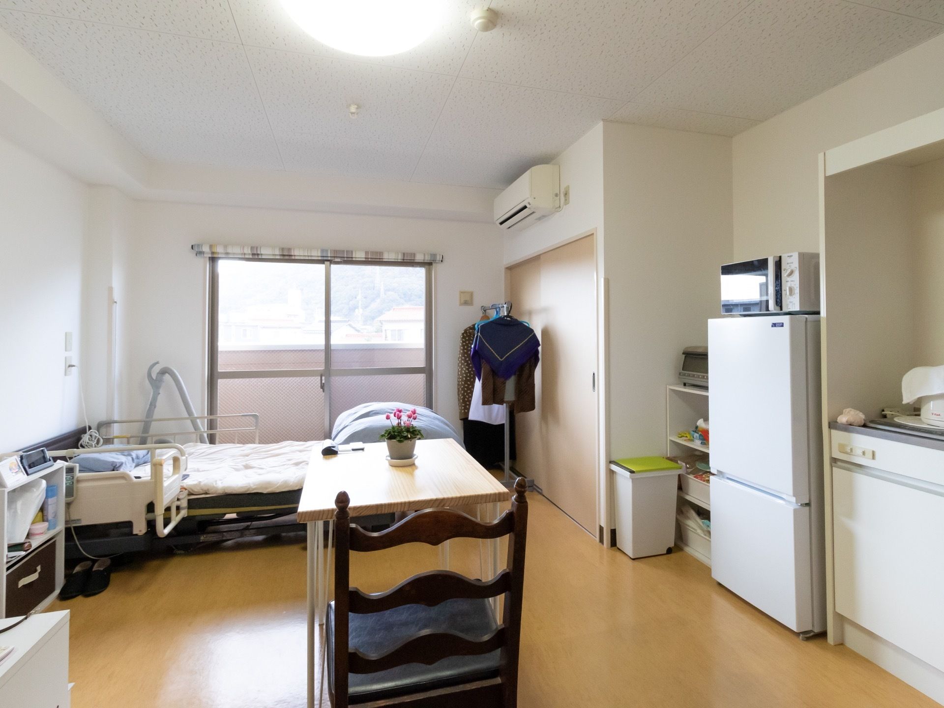 居室に掃き出し窓があり、窓越しにバルコニーの壁が見える。居室の中央部に、青い座面の椅子とテーブルがある。