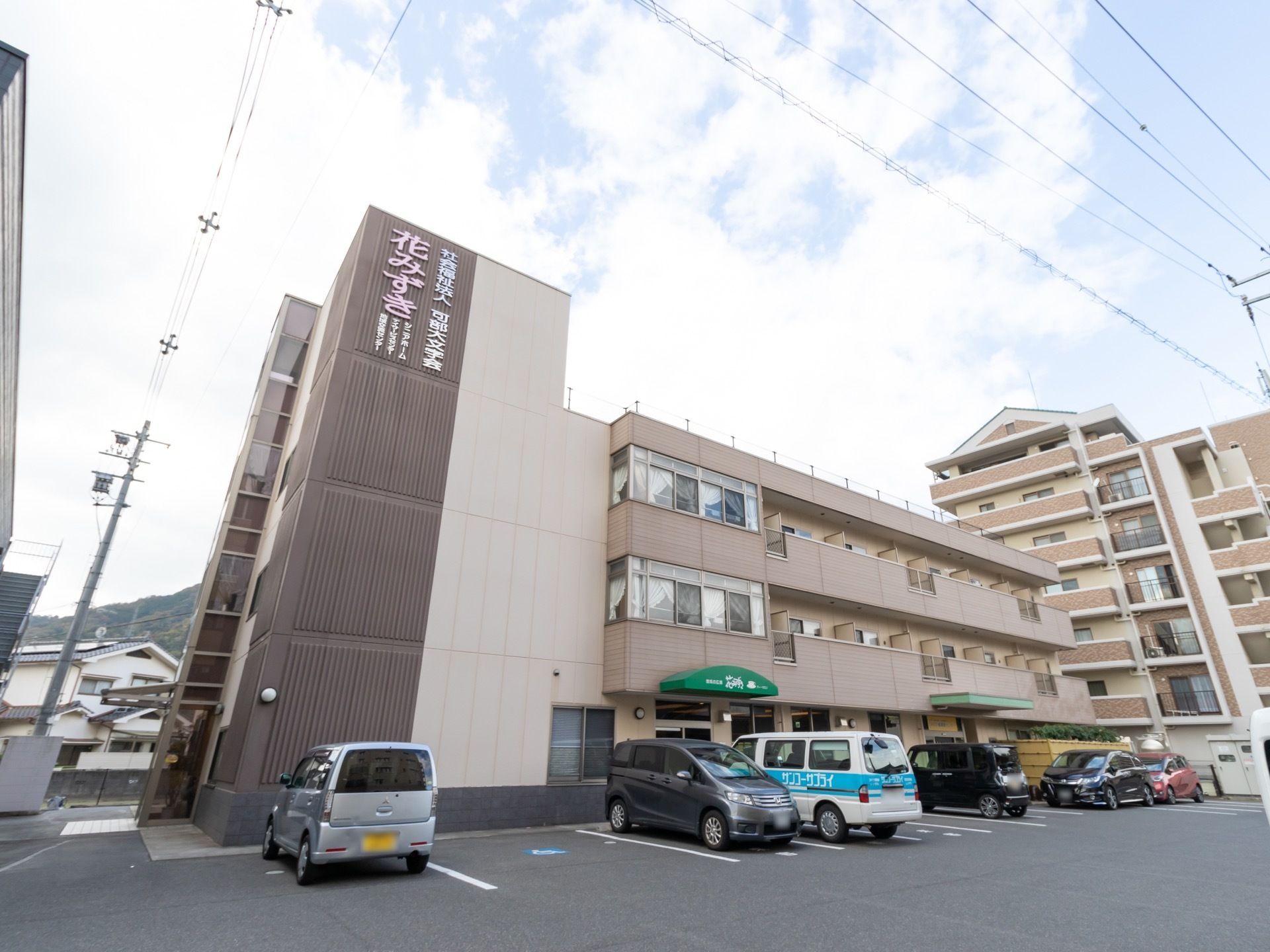 落ち着いた色調の外壁を持つ３階建ての施設である。建物前の敷地内に、白線で仕切られた多くの駐車スペースがある。