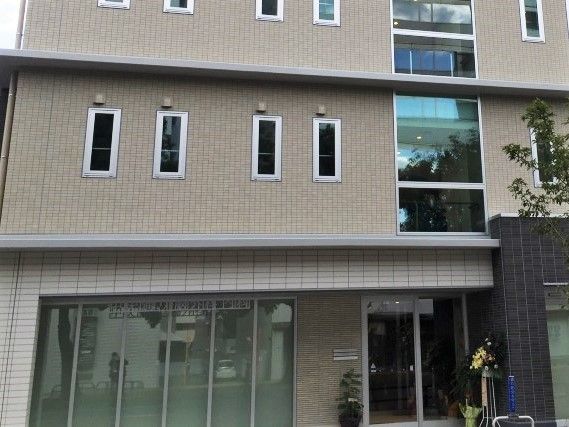 グレーの建物の一部。玄関周辺には観葉植物が置かれ、自動ドアが設置されている。