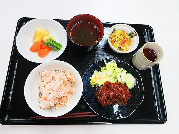温野菜の盛り合わせやサラダが添えられたハンバーグ、オレンジのご飯、お茶やスープ、野菜の和え物がある。