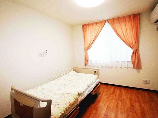 レースカーテンとカーテンが付いた大きな窓があるフローリングの部屋にはベッドや緊急通報装置もある。