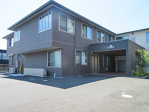 広い駐車場が付いた2階建ての建物はエル字型で玄関には住宅名が書かれた屋根も付いている。