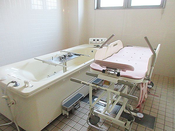 ピンクのストレッチャータイプのリフトが浴槽タイプの機械のそばに置かれている。大きな窓もある。