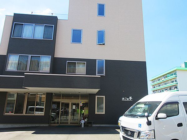 車が駐車場に止められている。ベージュとダークグレーの建物の玄関には車寄せが付いている。