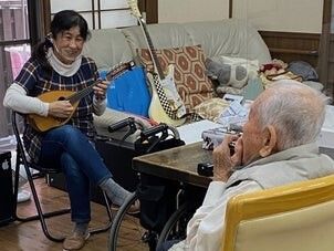 マンダリンを引いているジーンズをはいた女性とハーモニカを吹いている高齢男性が向かい合わせに座っている。