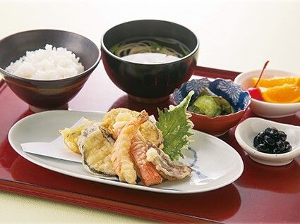 トレーの上には白いご飯や吸い物、天ぷらの盛り合わせ、野菜のおかず、フルーツなどが置かれている。
