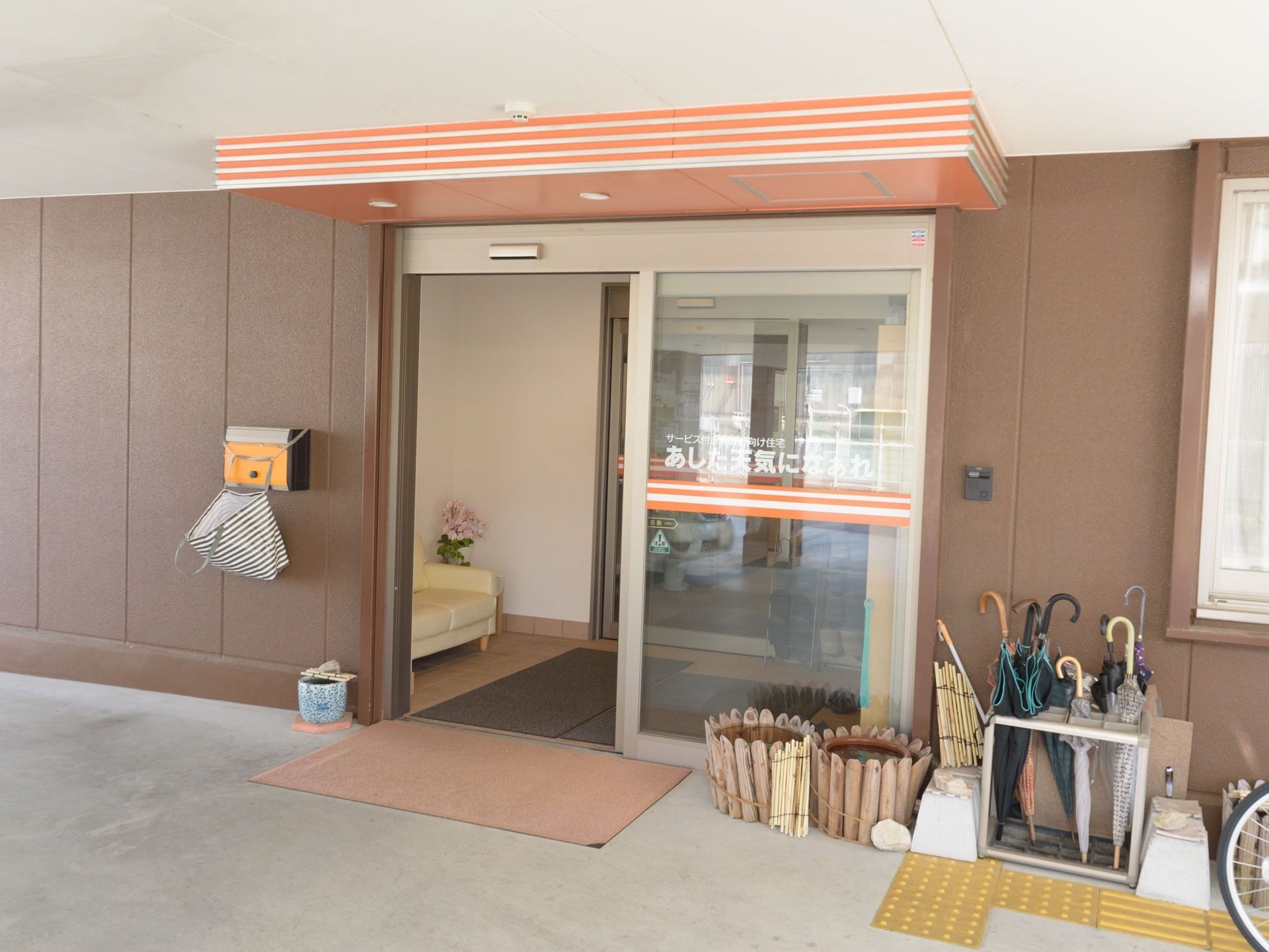 ガラス張りの自動扉の玄関ドア。玄関扉には施設名が大きく書かれている。足元には玄関マットと点字ブロックが設置されている。