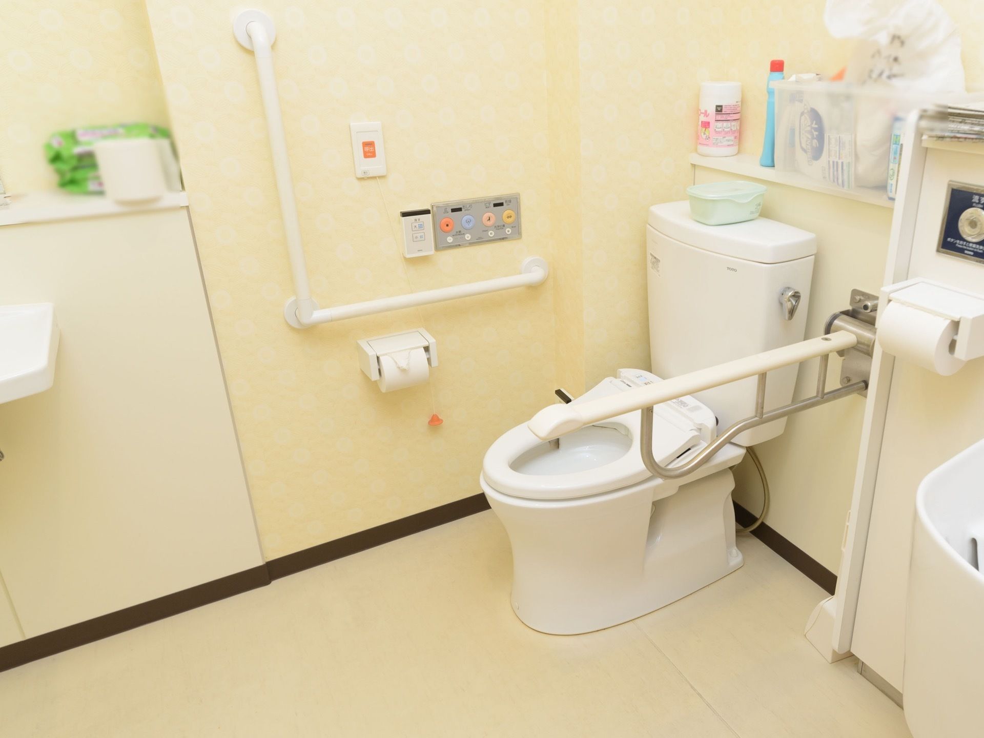 クリーム色の明るい壁紙の広いトイレがある。可動式のひじ掛けと手すりが設置されている。洗面台が側に用意されている。