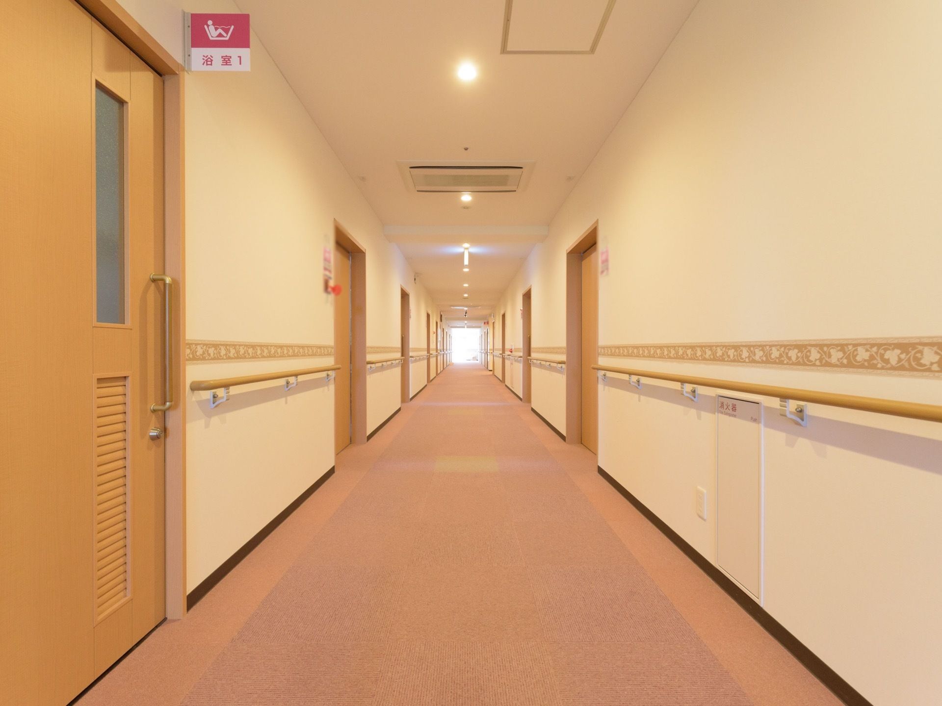 明るいベージュの床と落ち着いた照明器具の廊下がある。両側には居室の扉が並び、壁には手すりが設置されている。突き当りに大きな窓がある。