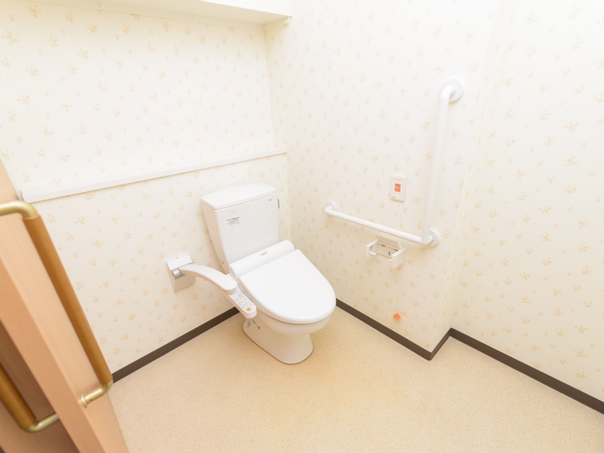 白い壁紙に囲まれた広いトイレがある。壁には縦横方面に手すりが設置されている。温水洗浄便座が備わっている。