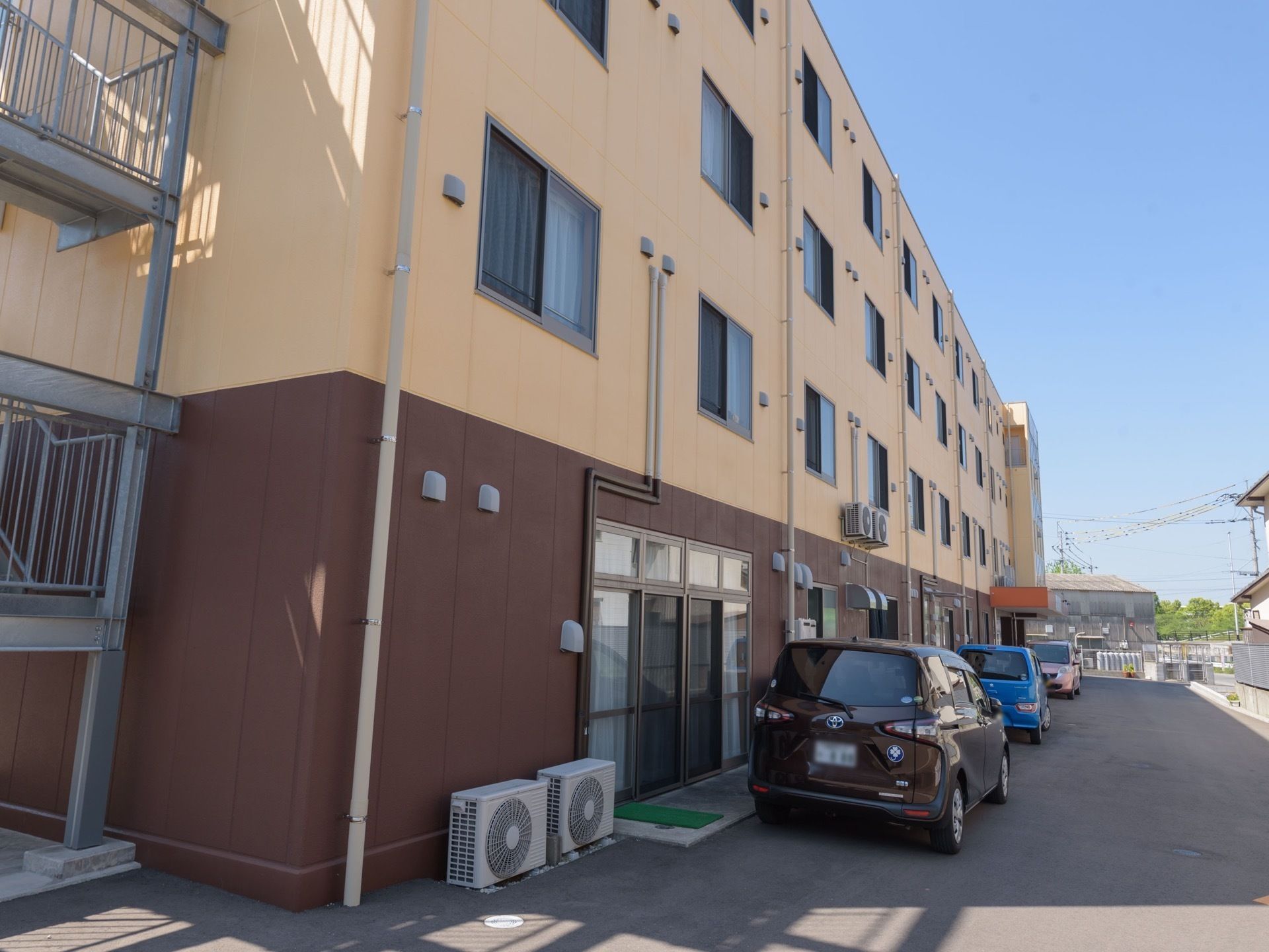 １階部分が茶色、上部分が黄色の４階建ての大きな建物がある。建物に沿って車が数台停まっている。建物の端には非常階段がある。