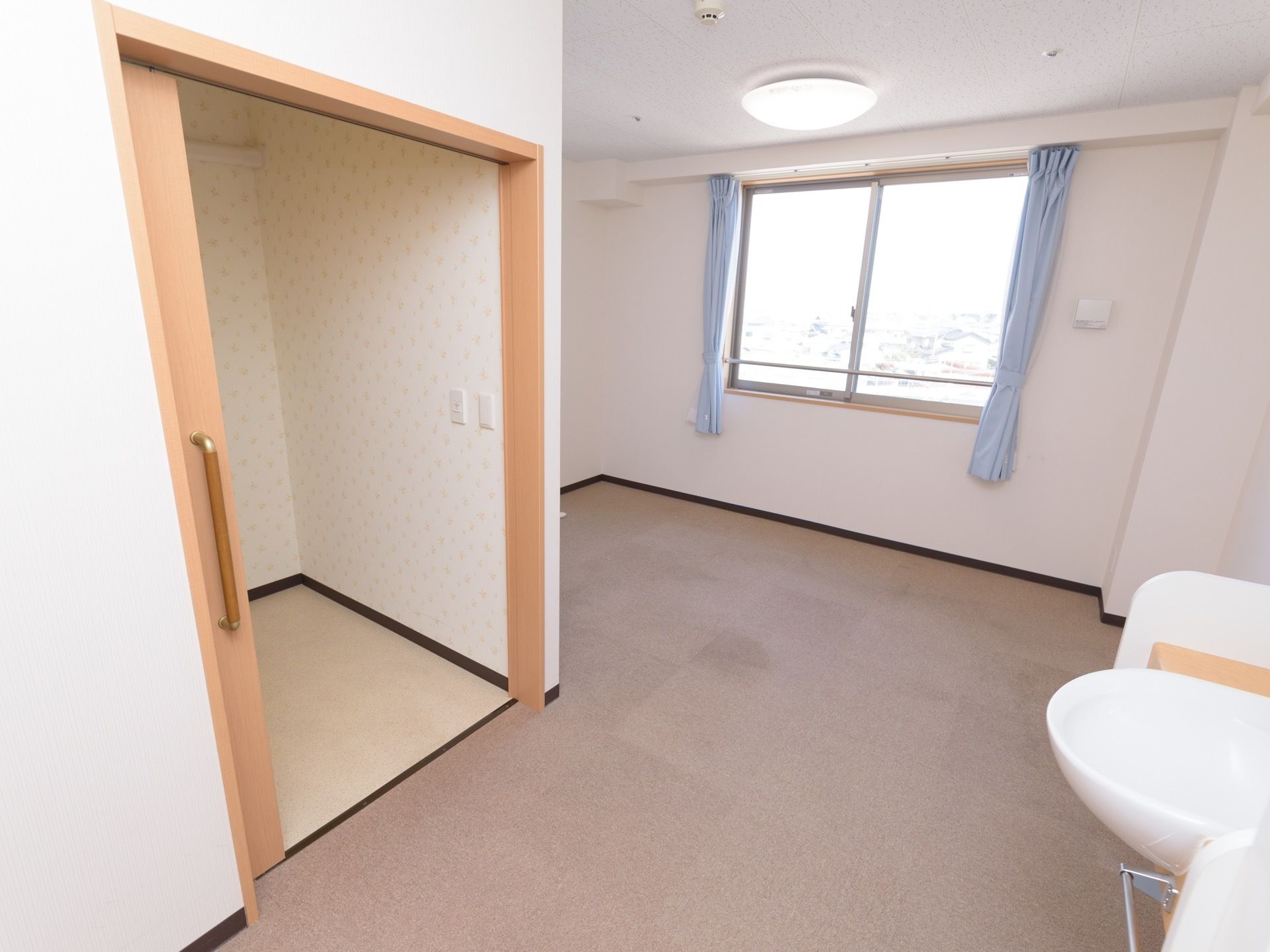 水色のカーテンが掛かっている窓のある居室。入り口付近にはトイレと洗面台がある。床はカーペットになっている。