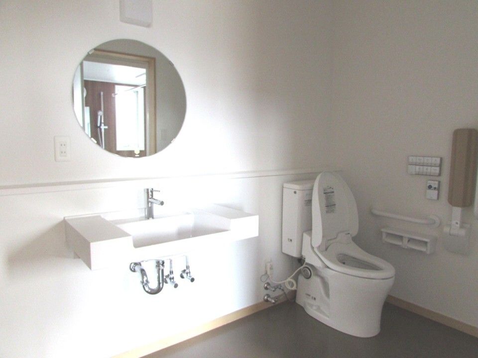丸い鏡や手洗い場が付いたシンプルなトイレは白を基調としていて、手すりも複数付いている。