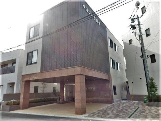 大きな車寄せがある3階建ての建物。ブラウン系で自動ドアの周りには木も植えられている。