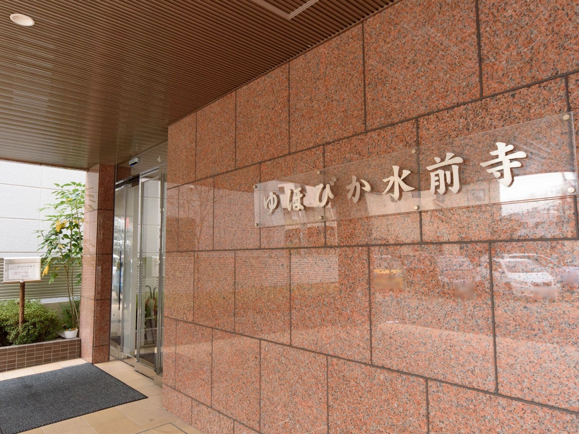 大きな屋根が設置されている玄関で、横には花壇が設けられている。外壁には、施設名が書かれた表札が取り付けられている。