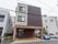 ブラウンを基調とした３階建てのスタイリッシュな外観の建物。横には駐車スペースが設けられており、複数台の車が停められている。